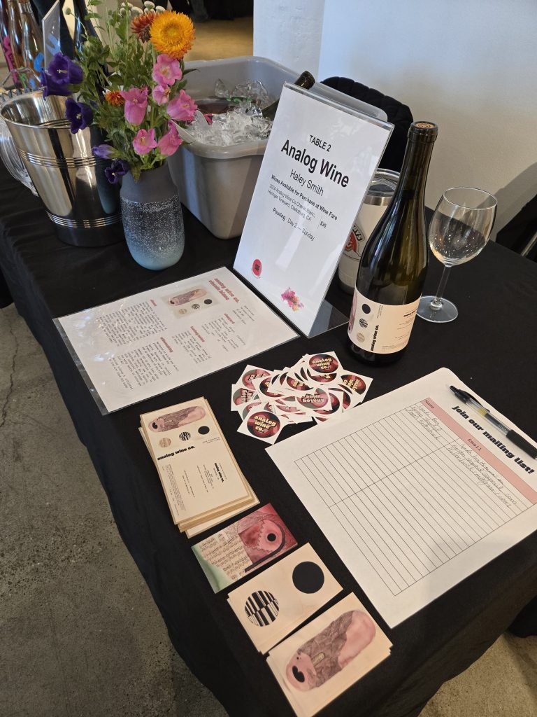 a table at a wine festival is covered with stickers, a sign up sheet, business cards, and wine bottles with the analog wine co branding